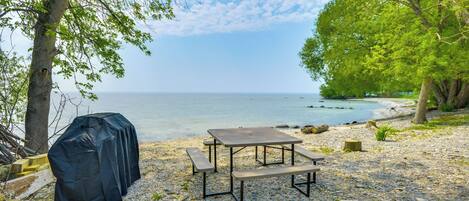 Ein Picknicktisch und ein Grill an einem kiesigen Strand mit Bäumen und Blick auf das Meer.