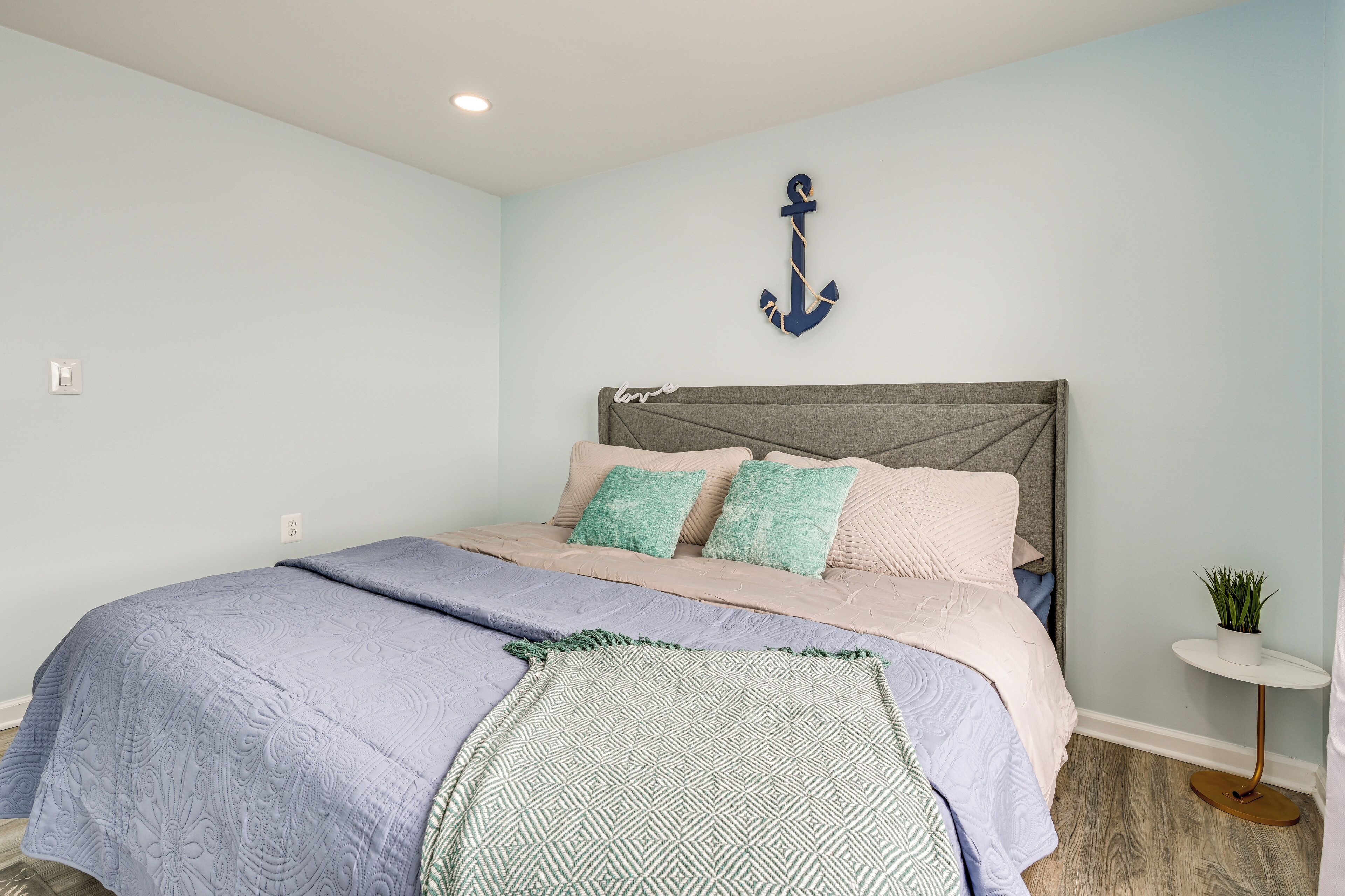 A bedroom with a bed, a headboard, a wall-mounted anchor decoration, and a small side table with a plant.