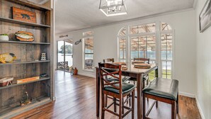 Un comedor con sillas y mesa de madera, un estante de madera con varios objetos, y ventanas amplias con vistas al exterior.