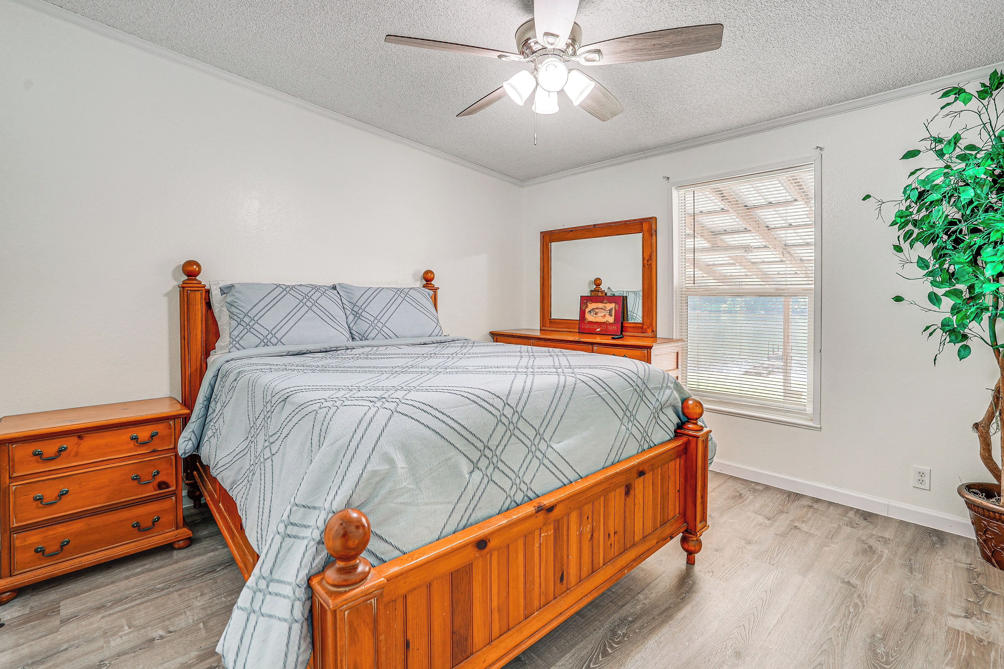 A bedroom with a wooden bed, a nightstand, a dresser, a ceiling fan, a window with blinds, and a potted plant.