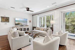 A spacious living room with a large flat-screen TV, a ceiling fan, and a view of greenery through large windows.