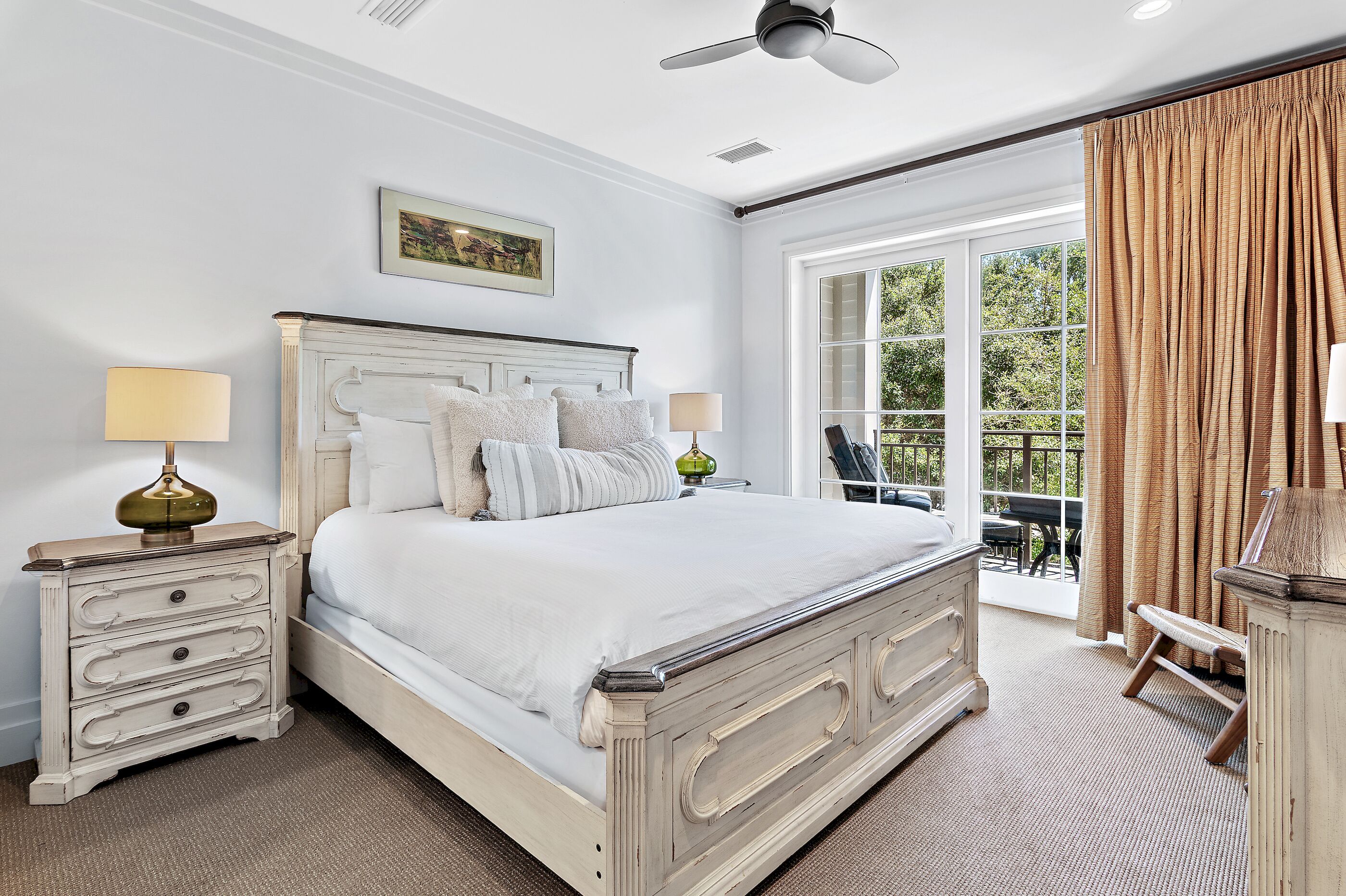 Une chambre à coucher comprenant un lit, des tables de chevet, un ventilateur de plafond, une fenêtre avec des rideaux et une vue sur la verdure à l’extérieur.