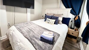 Een slaapkamer met een bed, een nachtkastje, een aan de muur bevestigde televisie en een raam met gordijnen.