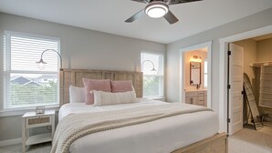 Une chambre avec un grand lit, un ventilateur de plafond, deux fenêtres équipées de stores, une table de chevet et un dressing.