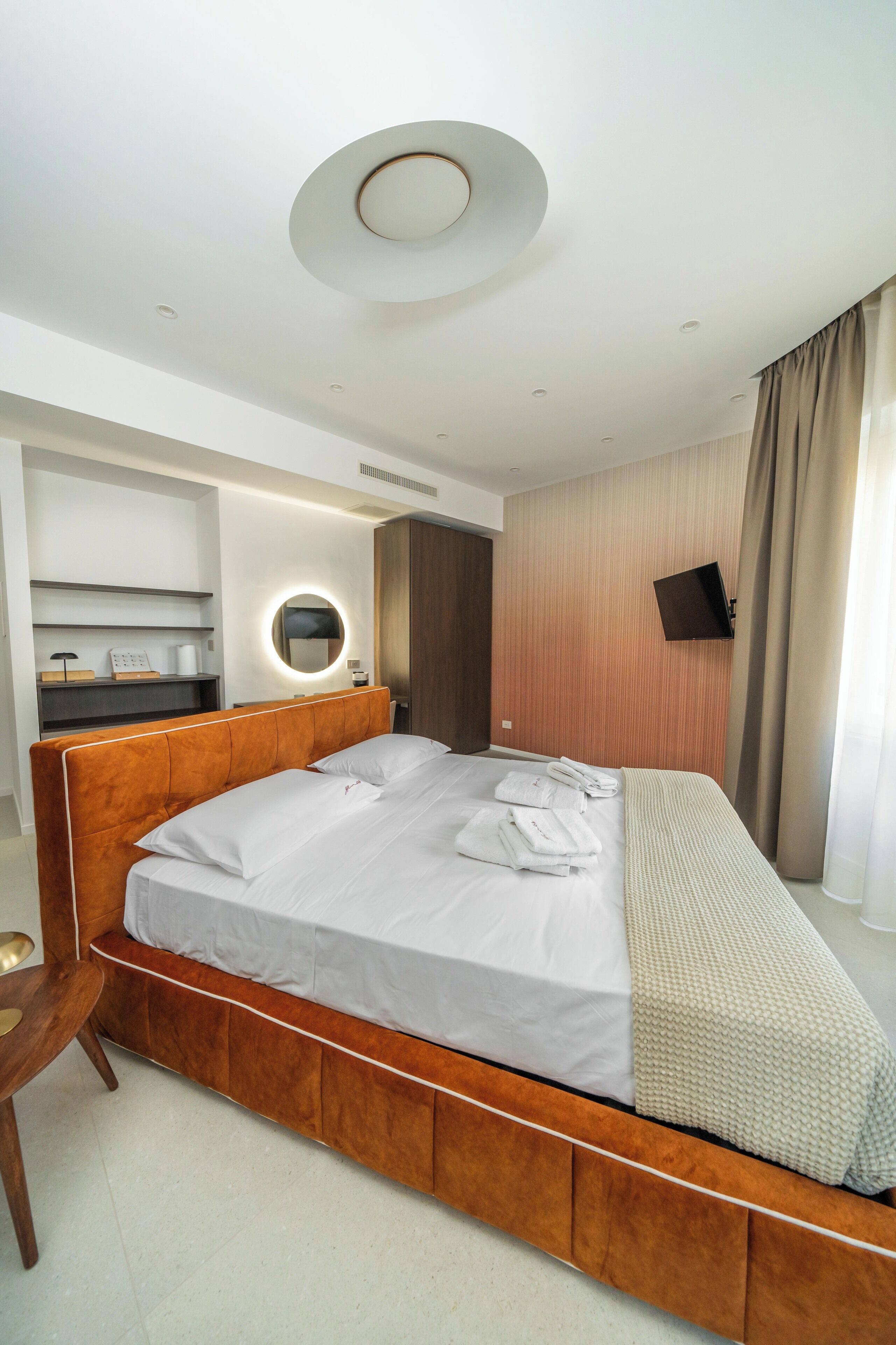 A modern hotel room with a wooden bed, a bedside table, a built-in shelf with a mirror, and a flat-screen TV mounted on the wall.