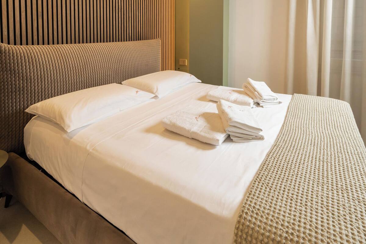A modern hotel room with a large bed, a wooden panel wall, and a view of the city through the window.