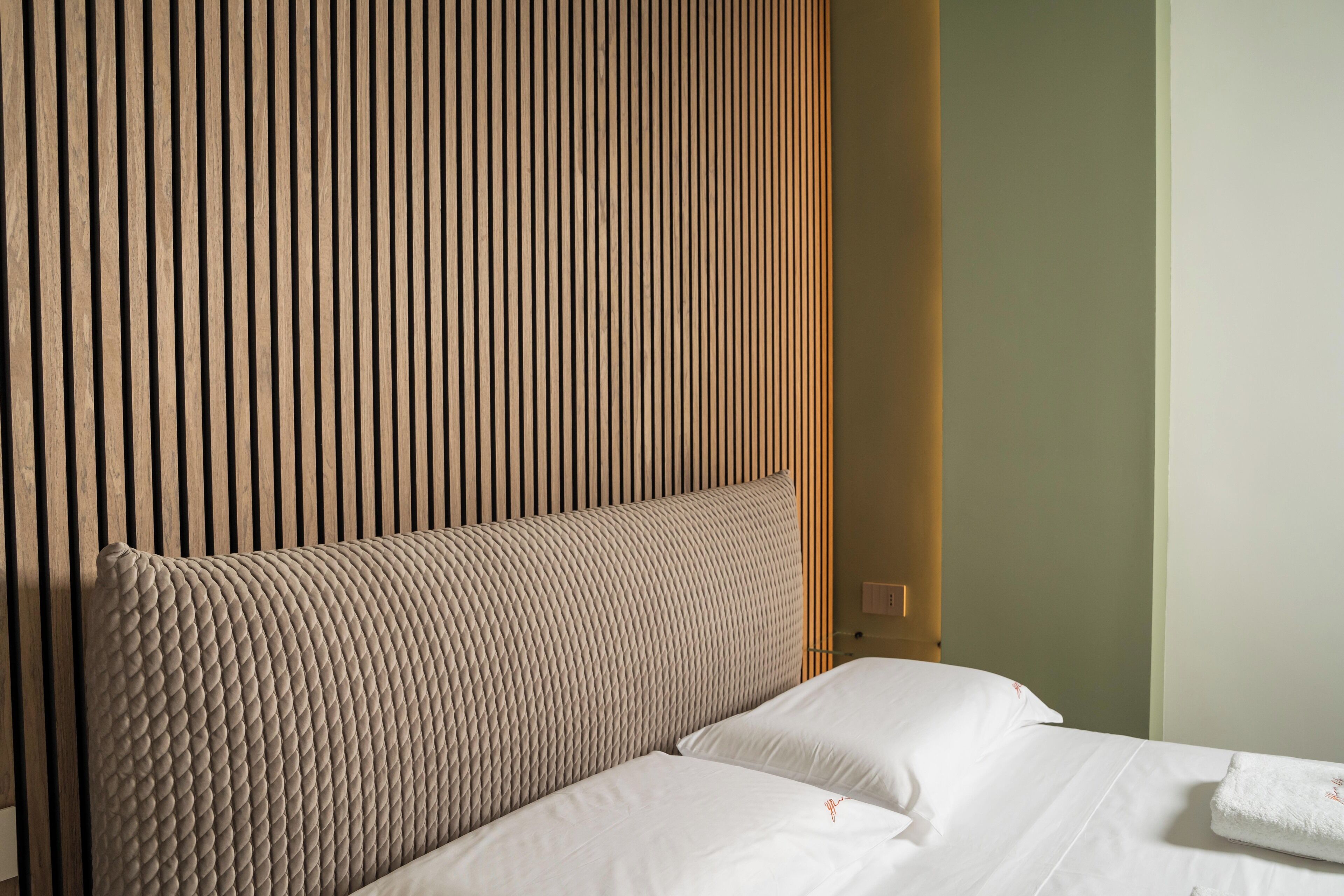 A hotel room with a wooden slat headboard, a bed with white linens, and a plain wall.