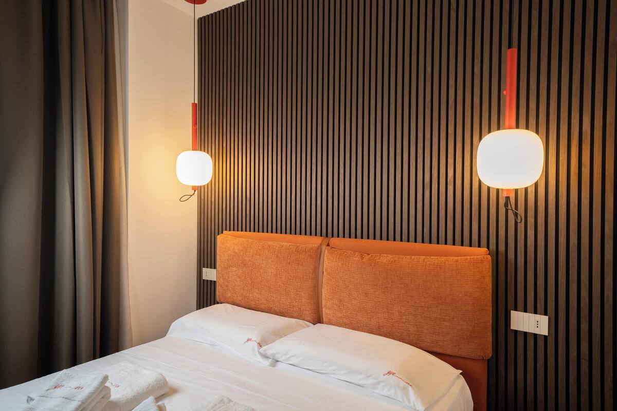 A hotel room with a bed, curtains, a headboard, and wall-mounted lighting.
