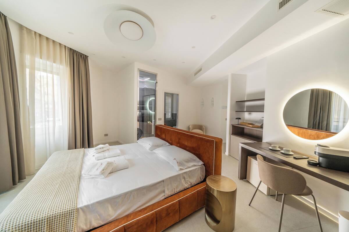 A modern hotel room with a wooden bed, a desk with a chair, a round mirror, and a large window with curtains.