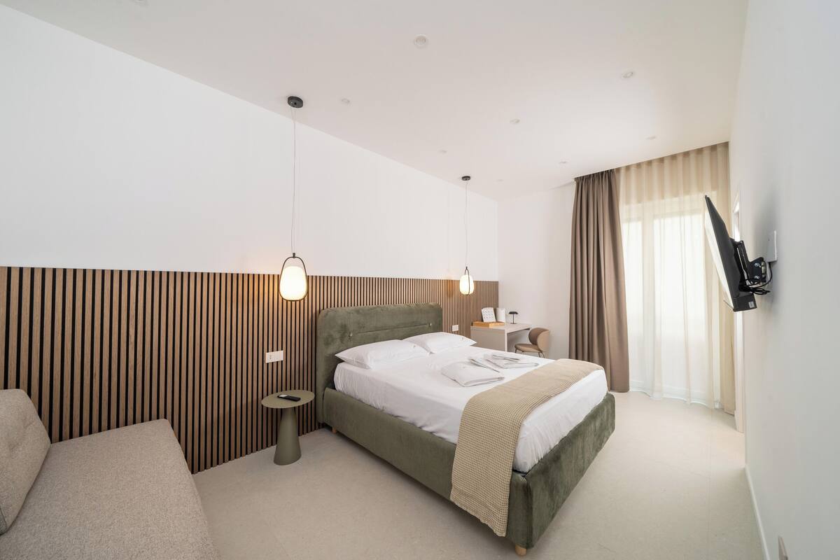 A modern hotel room with a large bed, a small bedside table, a sofa, a TV mounted on the wall, and a window with curtains.