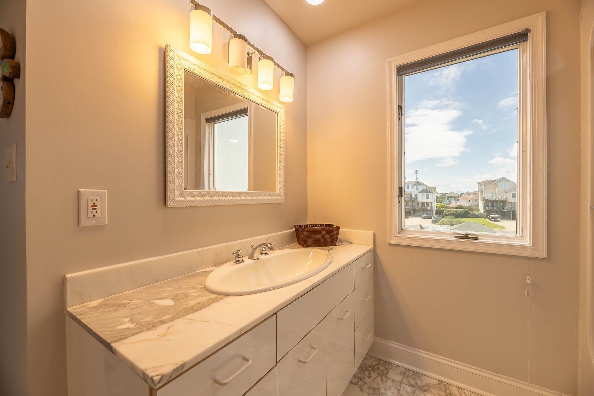 Une salle de bain avec un lavabo en marbre, un grand miroir avec éclairage intégré, une fenêtre donnant sur des maisons et un panier posé sur le comptoir.