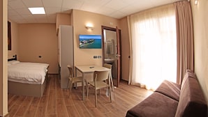 A hotel room with a bed, a desk with two chairs, a television mounted on the wall, a bathroom with a mirror, and a window with curtains. - Villa Canneto dep.Smeraldo (Sirmione)