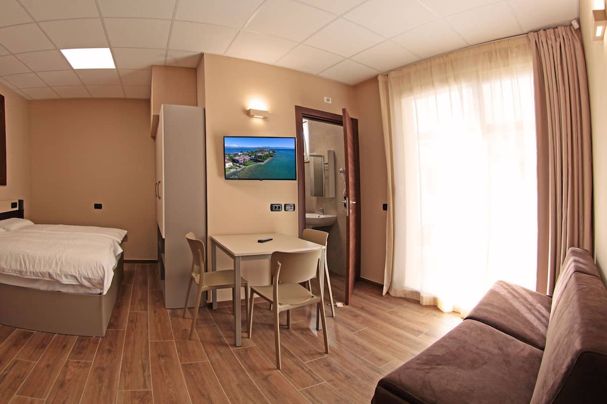 A hotel room with a bed, a desk with two chairs, a television mounted on the wall, a bathroom with a mirror, and a window with curtains.