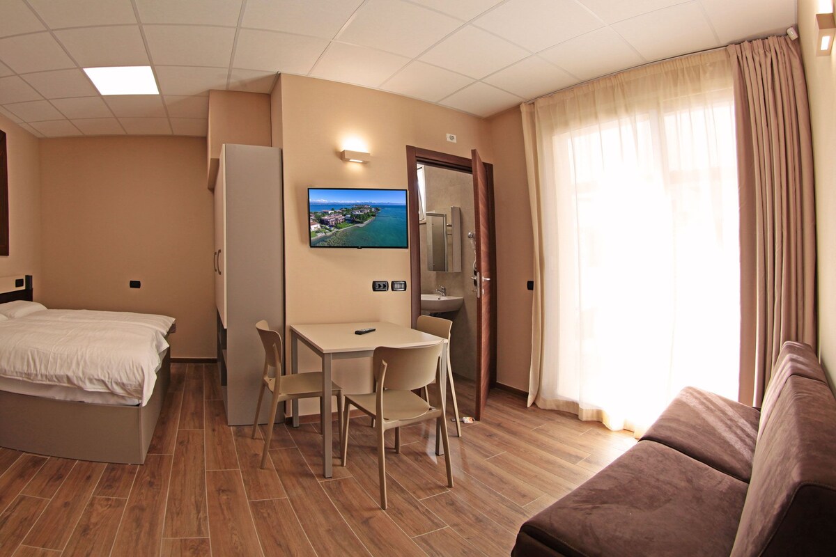 Habitación de hotel con cama, escritorio con dos sillas, televisor montado en la pared, baño con espejo y ventana con cortinas.
