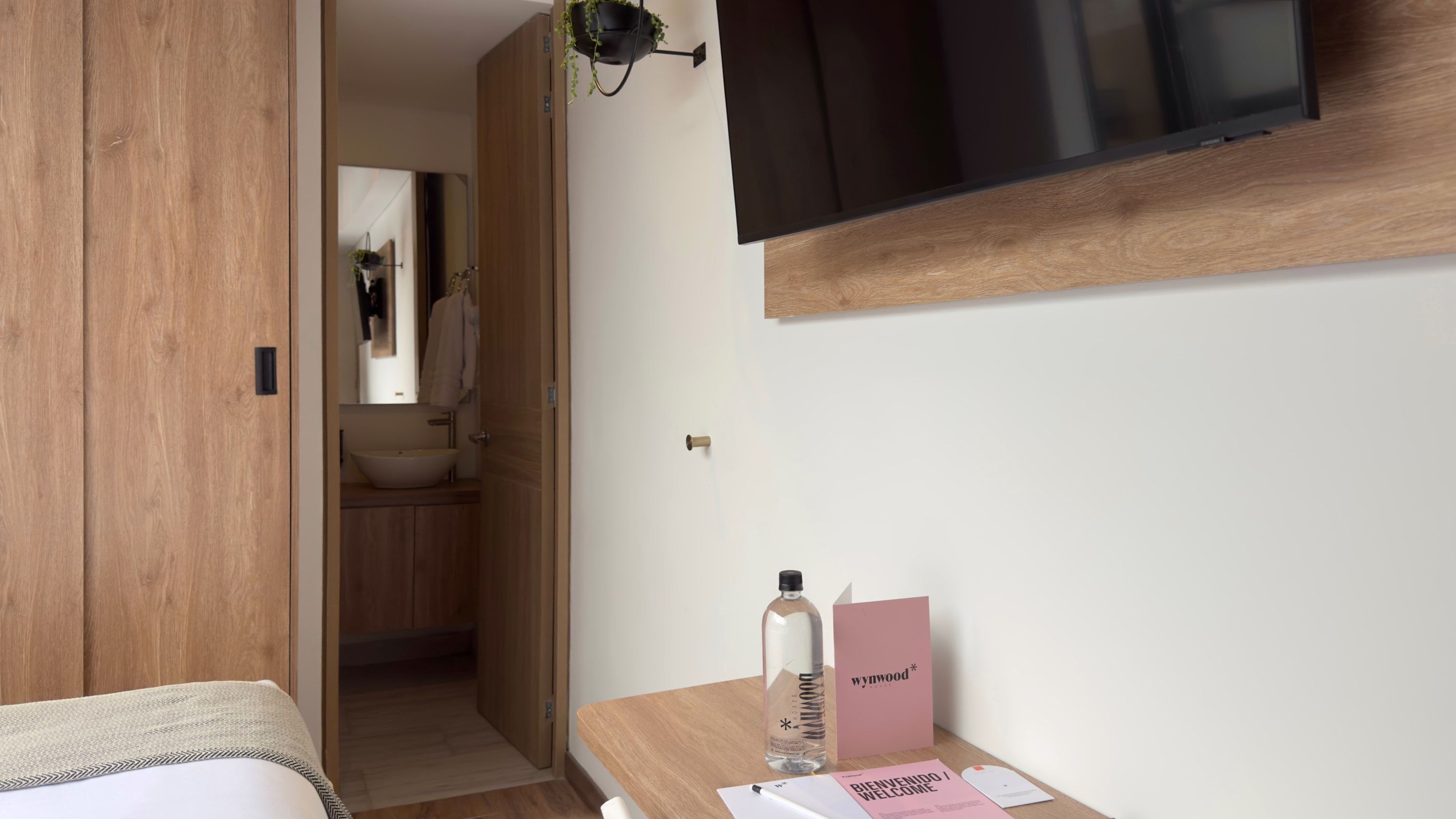 Une salle de bain moderne avec un meuble-lavabo en bois, un évier et un miroir. Une porte donne accès à une autre pièce, et un téléviseur est fixé au mur au-dessus du meuble-lavabo.
