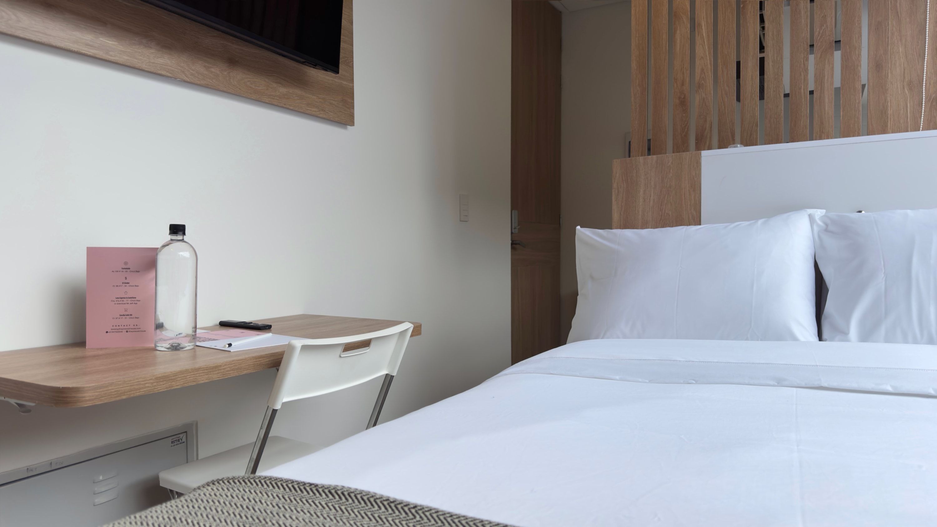 Ein Hotelzimmer mit Bett, Schreibtisch mit Stuhl, einer Flasche und einer rosa Karte.