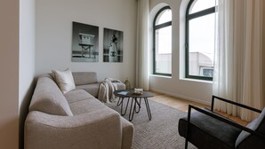 A modern living room with a beige sofa, a black armchair, and a small round table. There are two large windows with curtains, and artwork is displayed on the wall. - Stunning Seafront Apartment With Modern Comforts and Dike Views (Ostend)