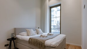 A modern bedroom with a large bed, a small bedside table, and a window with a view of buildings. - Stunning Seafront Apartment With Modern Comforts and Dike Views (Ostend)