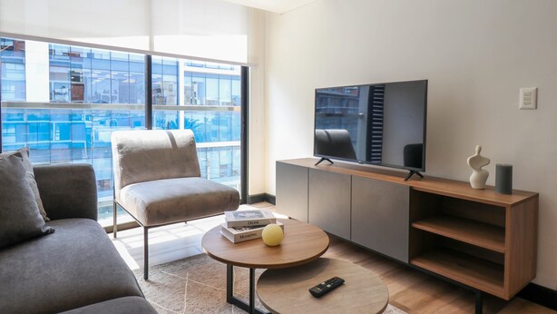 A modern living room with a sofa, armchair, and a coffee table with books and a remote control. A TV is mounted on a wooden console, and a window offers a view of the city. - w Resplendent 2BR w Great Balcony in San Isidro (Lima)