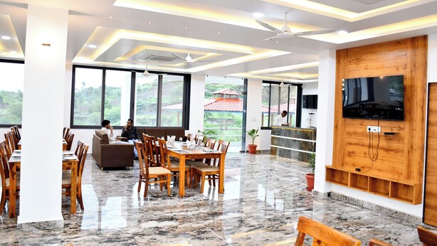 A spacious indoor area with a dining table, chairs, a sofa, a TV mounted on a wooden panel, and large windows offering a view of greenery outside. - Hotel RC Garh (Nathdwara)
