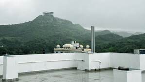 A rooftop terrace with a view of a hilltop structure, a white building with domes, and a tall chimney. - Hotel RC Garh (Nathdwara)