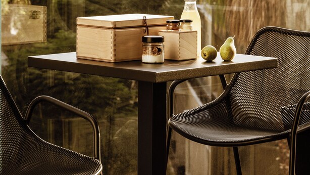 A balcony with a table set with boxes, a bottle, and two apples. There are two wicker chairs and a view of buildings and palm trees. - Likno Athens (Athens)