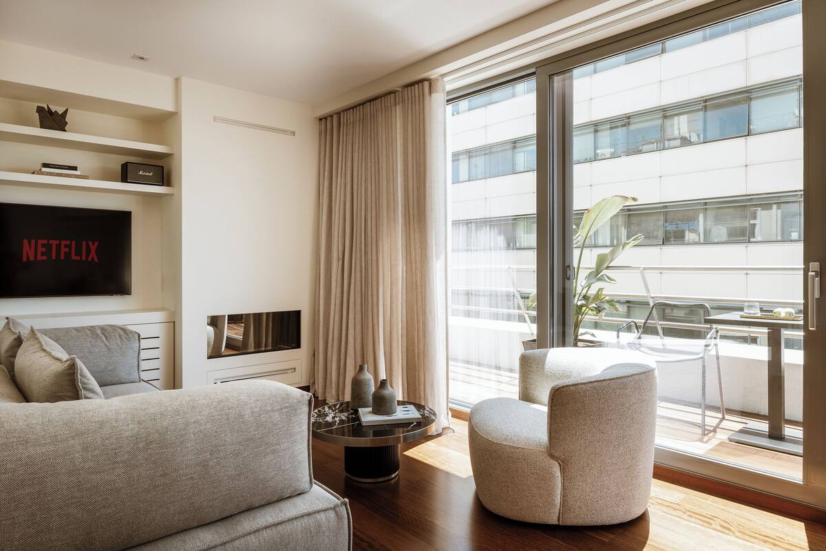 A modern living room with a sofa, armchair, and a coffee table. A flat-screen TV is mounted on the wall, and there is a balcony with a view of the city.