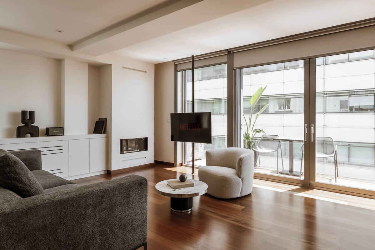 A modern living room with a flat-screen TV, a sofa, a coffee table, and a balcony with a plant.
