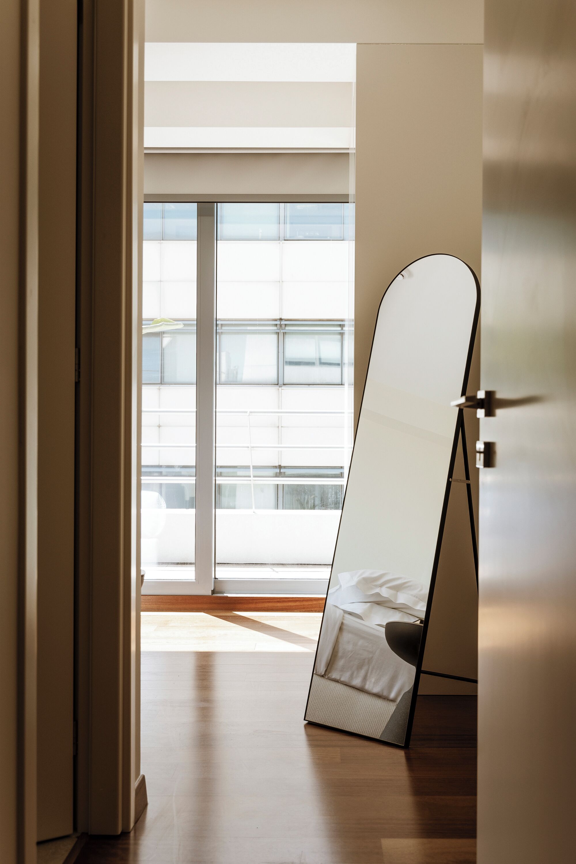 Una habitació amb una finestra gran, un llit i un mirall adossat a la paret.