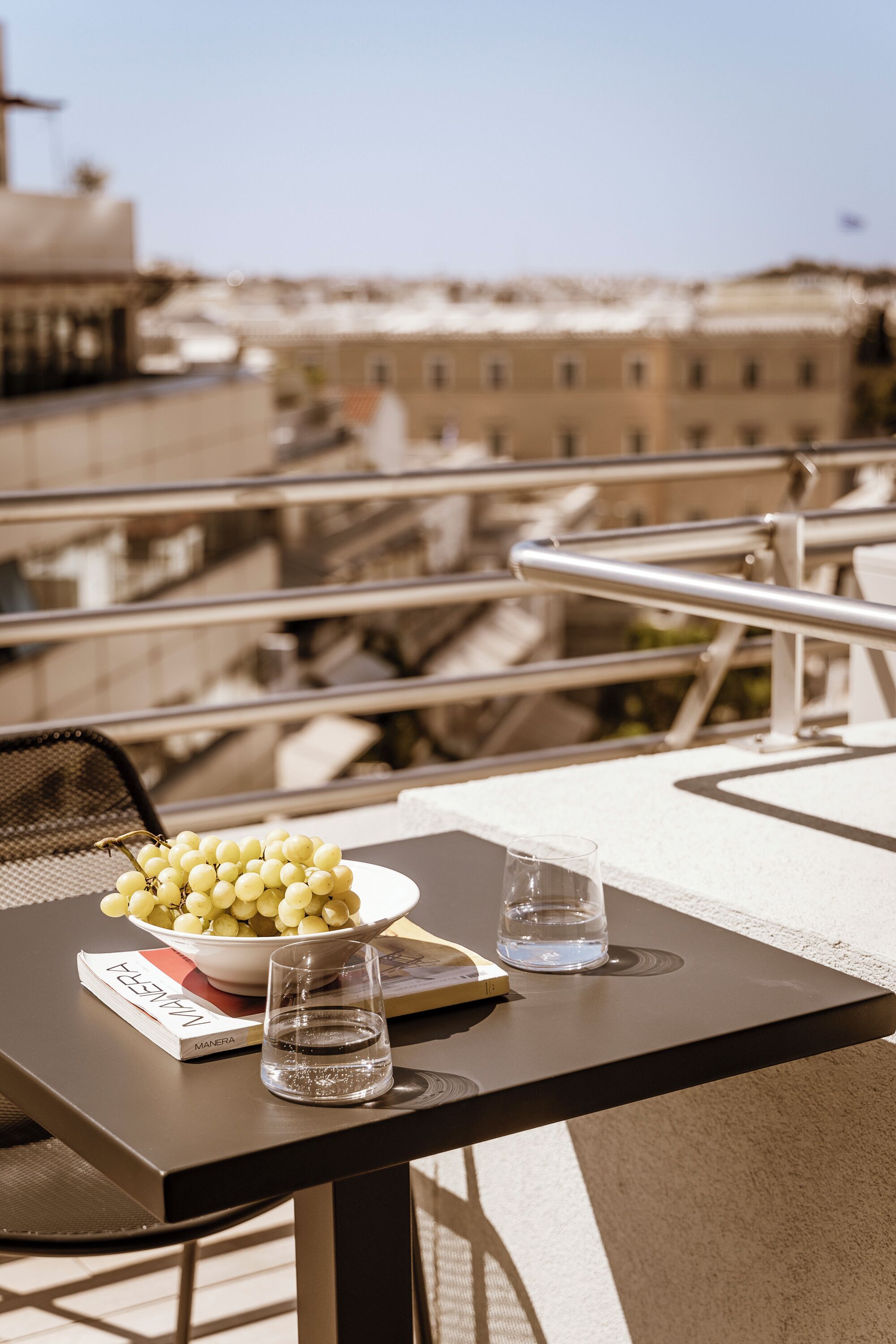 A table with a bowl of grapes, a glass of water, and a book titled "MANERA" on a balcony overlooking a cityscape.