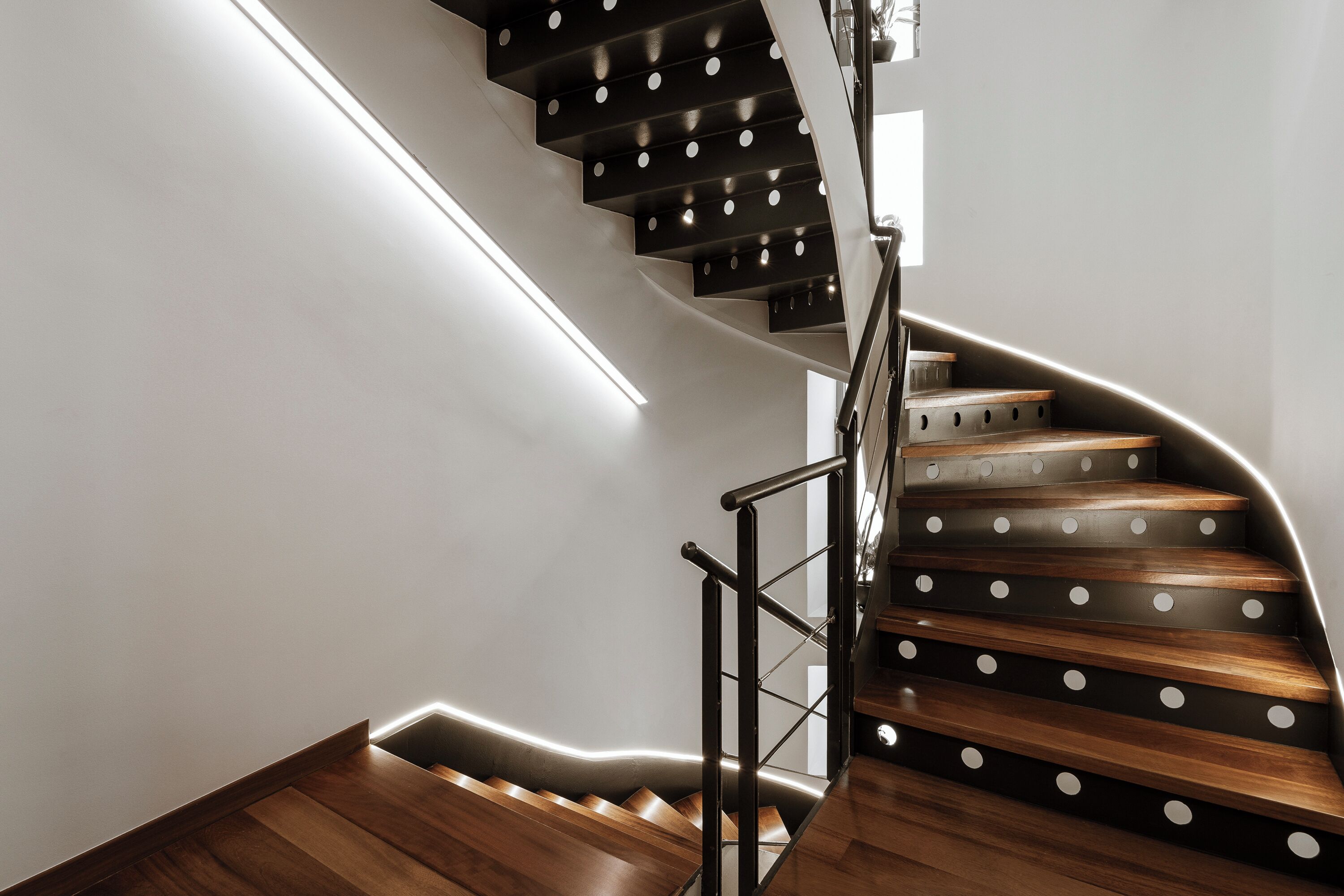 A modern spiral staircase with wooden steps and black railings, illuminated by under-stair lighting.
