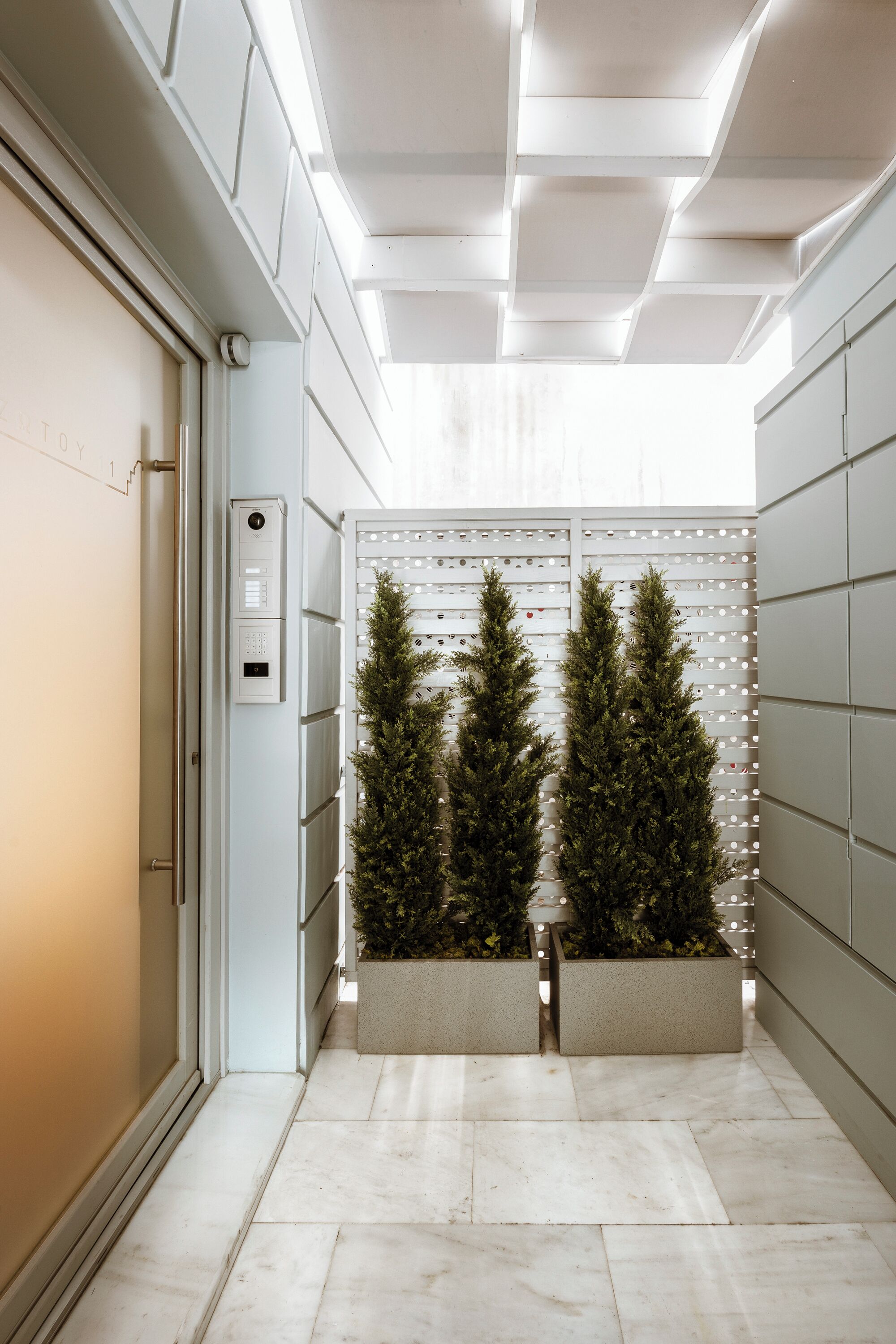 A hallway with a modern design, featuring a door with a control panel, a wall-mounted light fixture, and two potted evergreen trees.