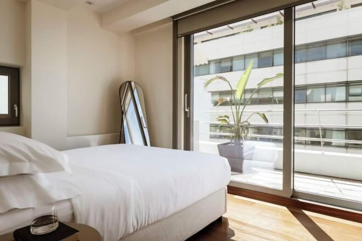 A bedroom with a bed, a bedside table, a potted plant, and a view of a building through a sliding glass door.