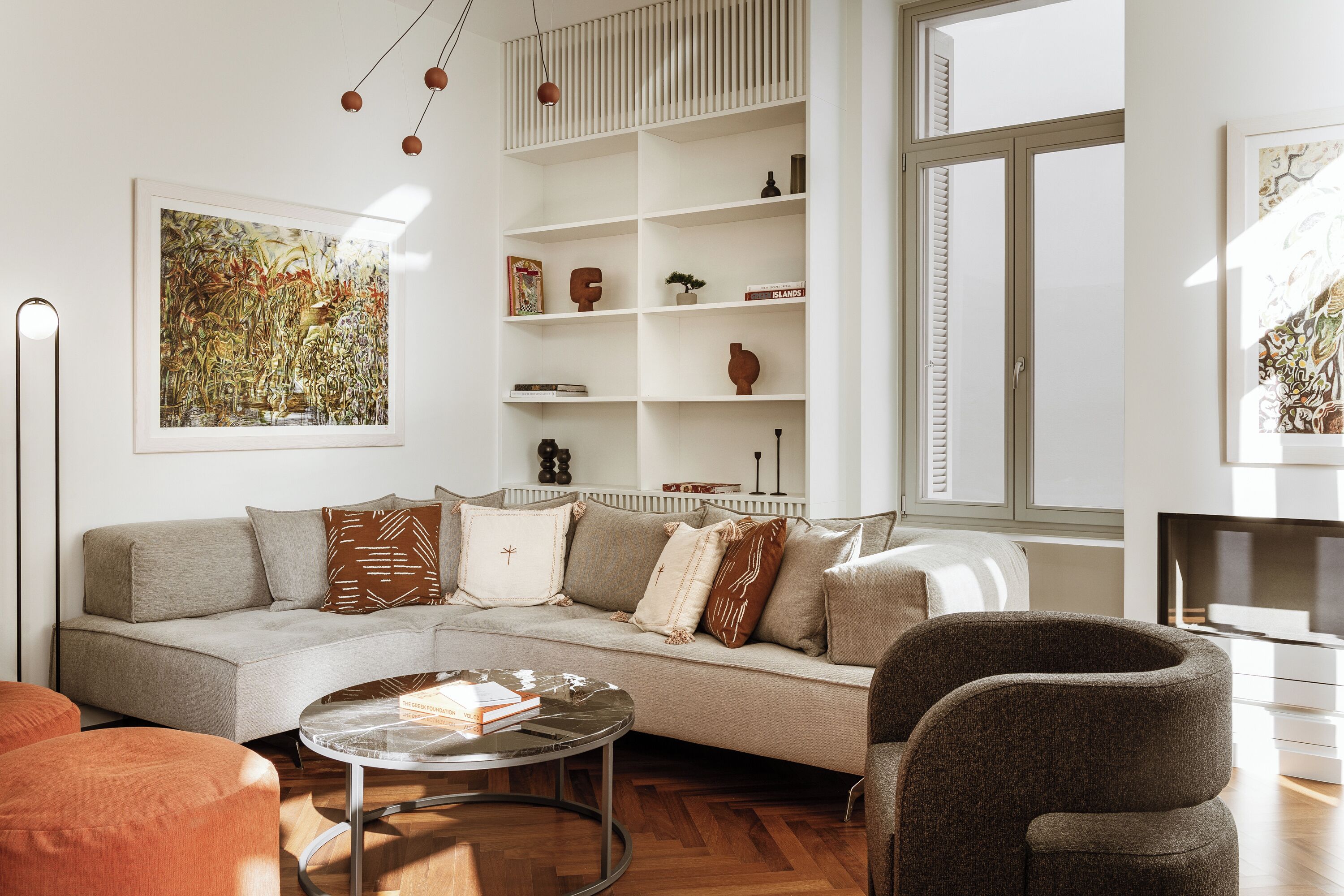 A modern living room with a sofa, coffee table, and armchairs. There is a bookshelf with decorative items and a painting on the wall.
