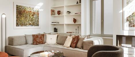 A modern living room with a sofa, coffee table, and armchairs. There is a bookshelf with decorative items and a painting on the wall.