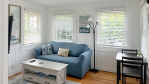 A room with a blue sofa, a white coffee table, a small table with a chair, and a floor lamp. There are two windows with blinds, a wall-mounted light, and a framed picture on the wall. - Billy's Beachside Bungalow - near Wallis Sands Beach (Rye)