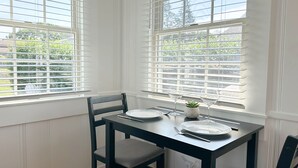 A dining area with a table set for two, a chair, and a window with blinds. - Billy's Beachside Bungalow - near Wallis Sands Beach (Rye)