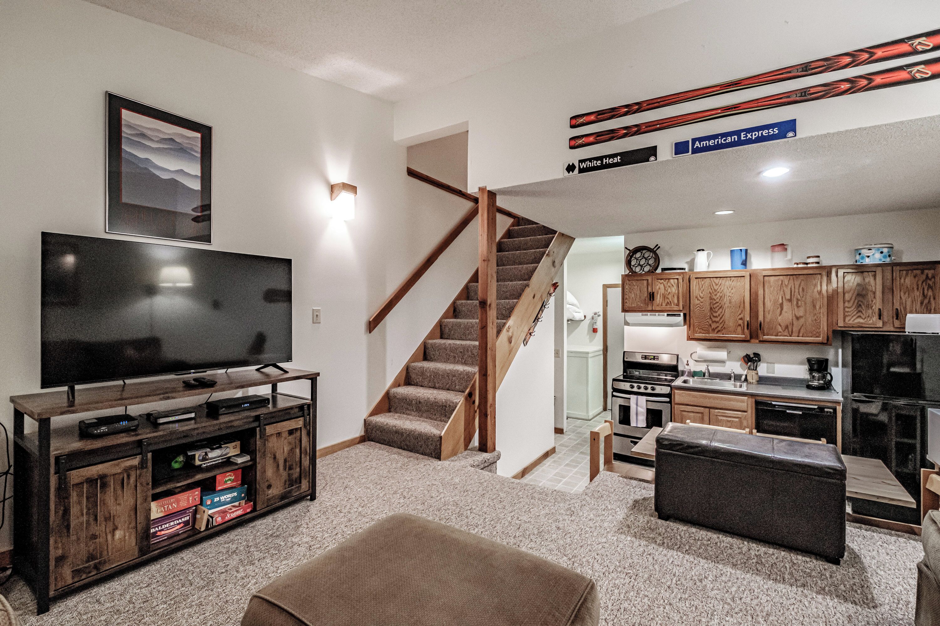 A modern living room with a flat-screen TV, wooden entertainment unit, and a staircase leading to a kitchen area.