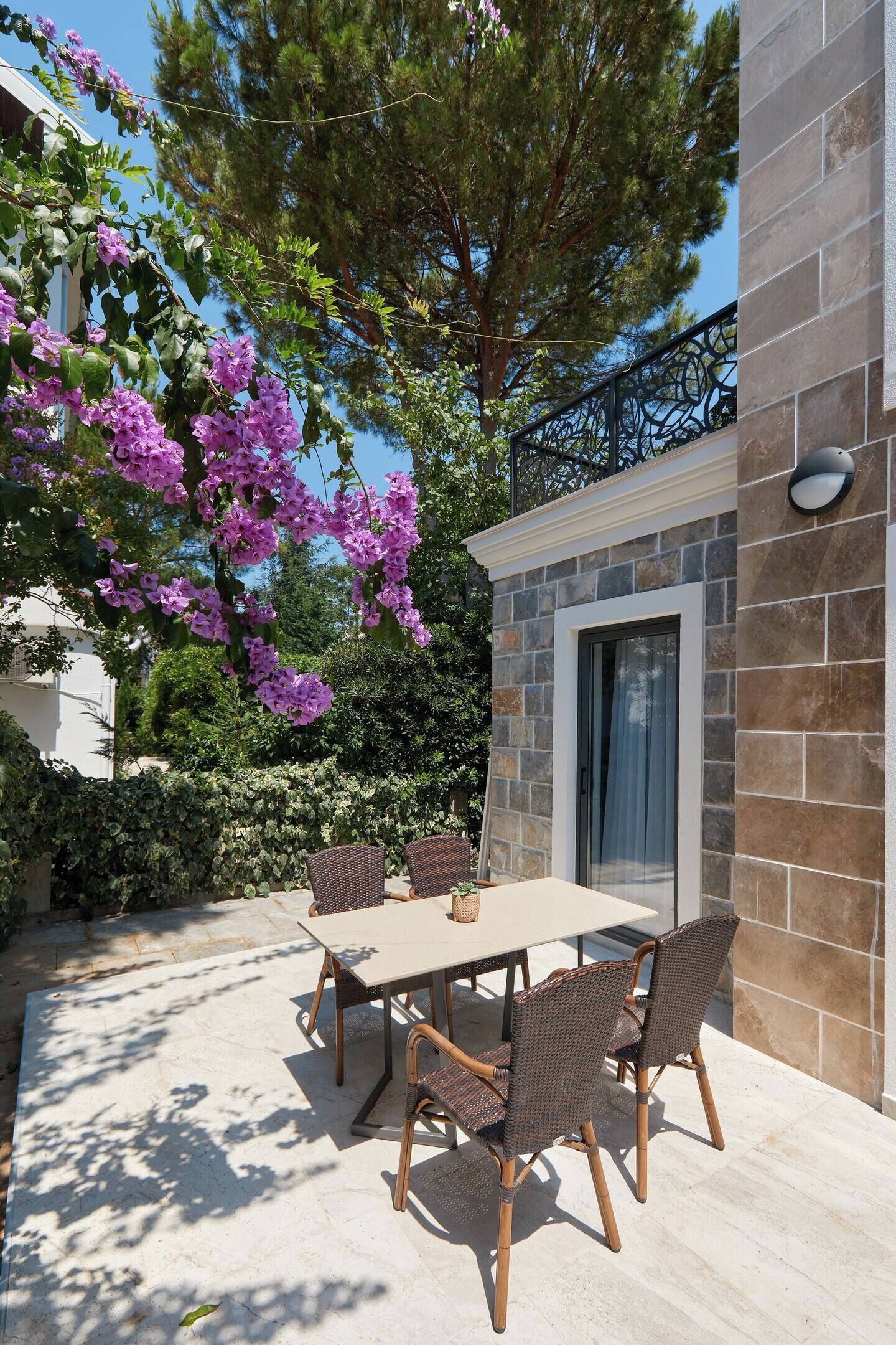 Eine Terrasse im Freien mit einer Steinmauer, einem Tisch mit Stühlen und violetten Blumen.