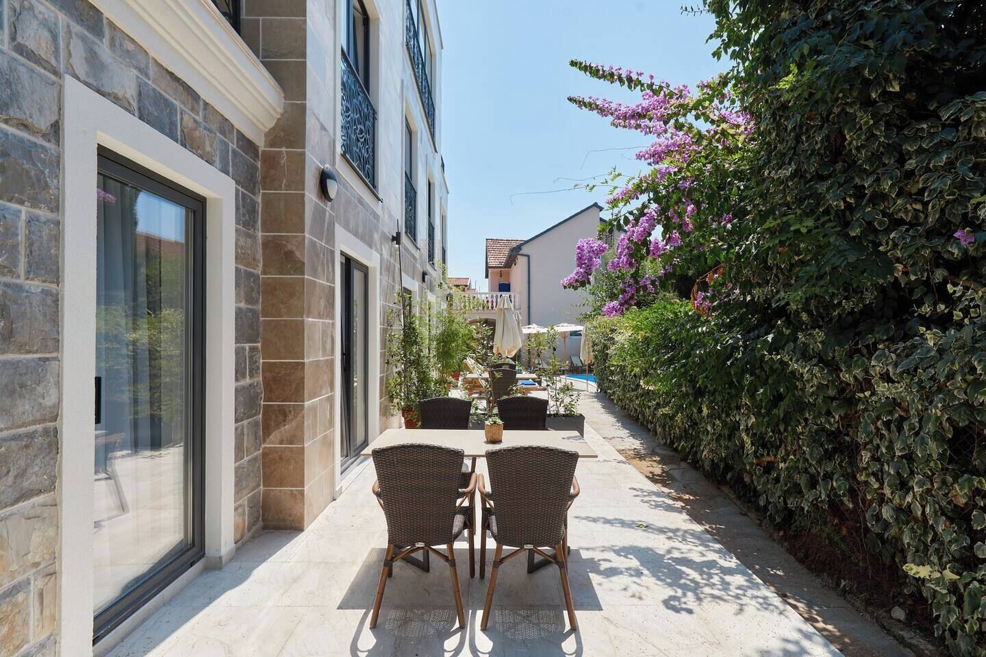 En smal udendørs terrasse med stenvægge, fletmøbler og udsigt til et poolområde og en bygning i baggrunden.