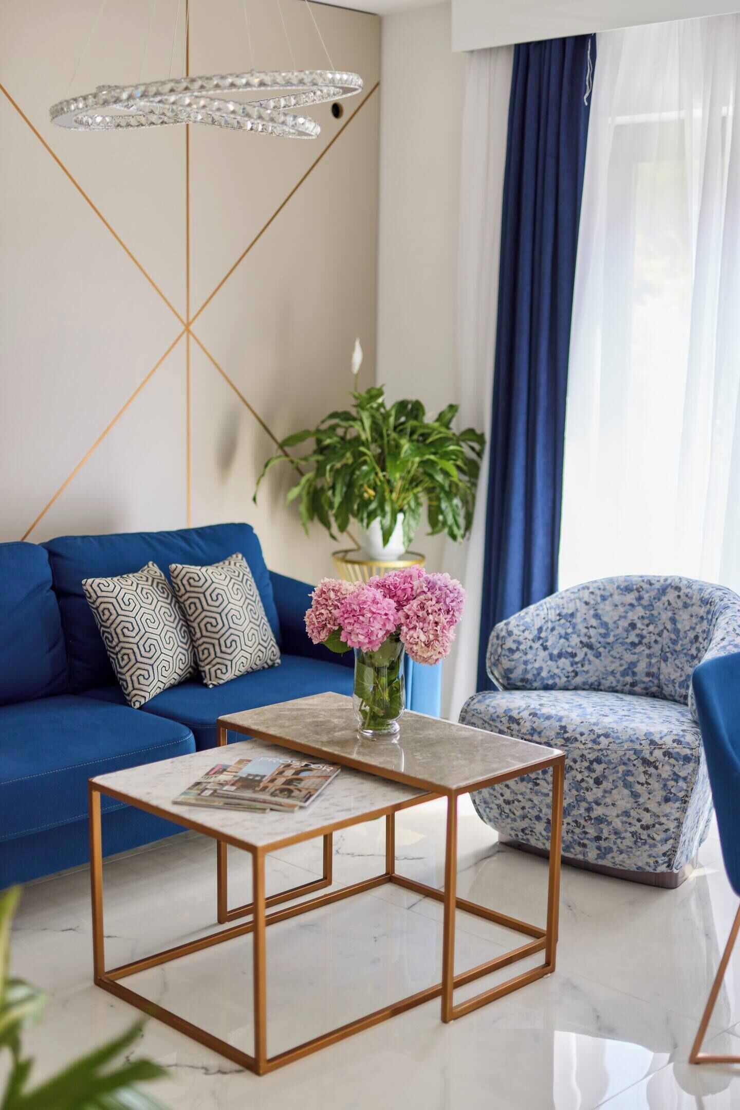 Ein modernes Wohnzimmer mit einem blauen Sofa, gemusterten Kissen, einem blumigen Sessel, einem Marmor-Couchtisch und einer hängenden Chandelieber.
