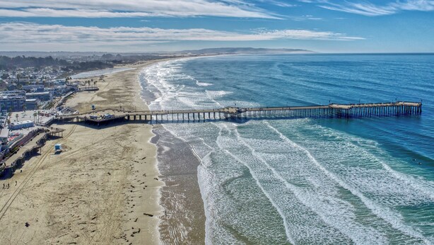 Una playa con un muelle que se adentra en el océano, una orilla arenosa y un horizonte urbano distante.