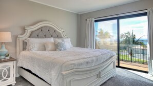 Una cama grande con cabecera capitonada, una mesita de noche con lámpara y vistas a un balcón con palmeras.
