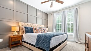 Un dormitorio con cabecera capitonada, cama con cubrecama acolchado, lámpara de noche, mesita de noche y ventana con cortinas.