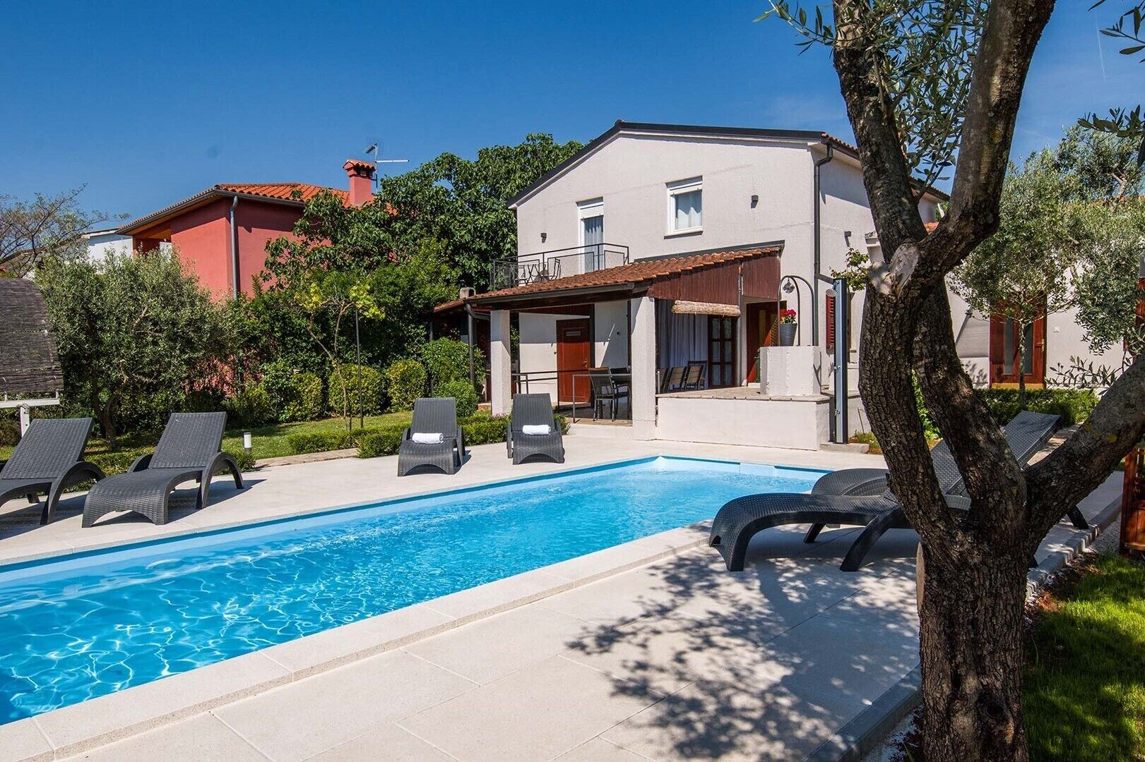 Une piscine avec des chaises longues, un bâtiment blanc doté d’un balcon et un arbre au premier plan.