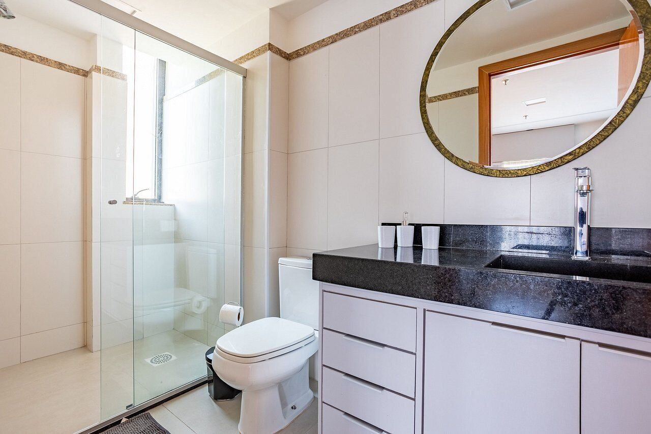 A modern bathroom with a glass-enclosed shower, a white toilet, a black countertop, and a round mirror with a gold frame.