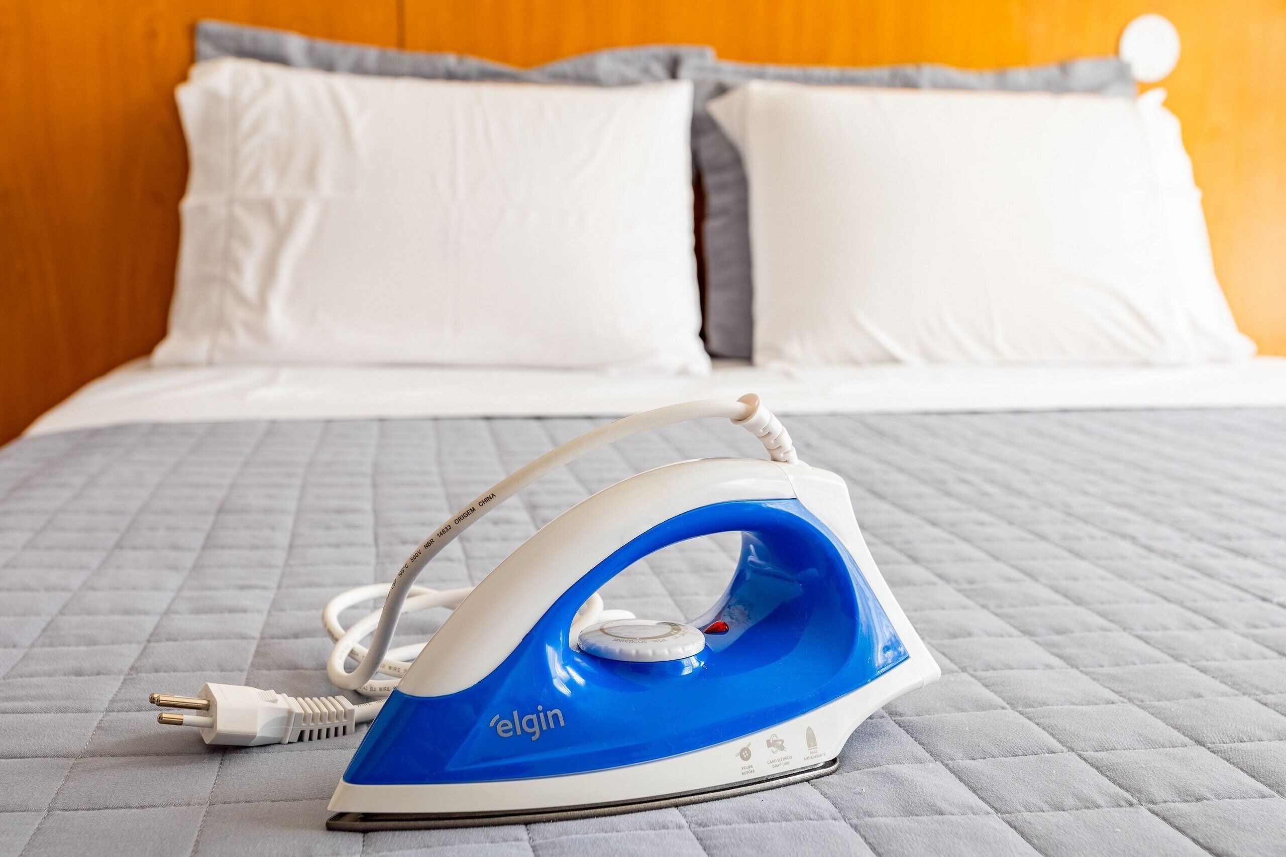 Um ferro da Elgin sobre uma cama com manta acolchoada e almofadas brancas.
