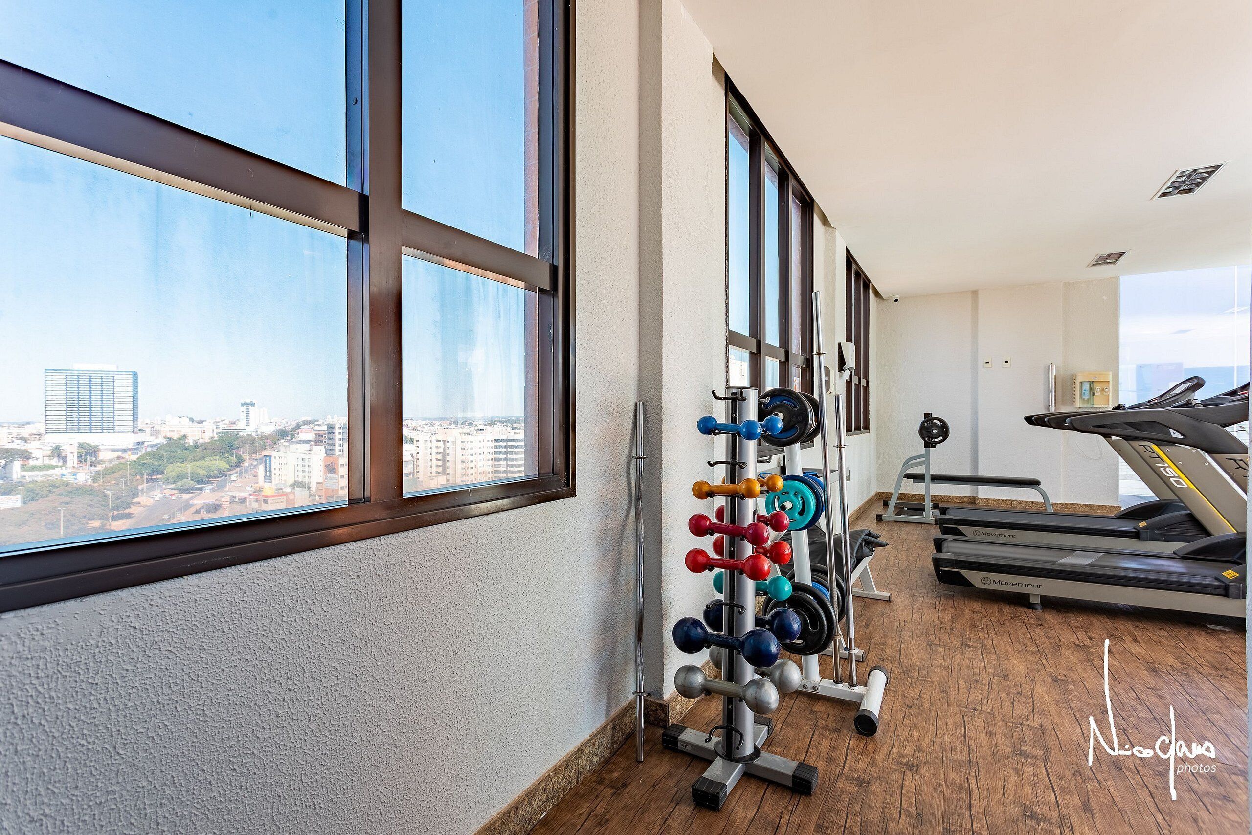 Un gimnasio con equipo de levantamiento de pesas y cintas de correr, ventanas amplias y vistas a la ciudad.