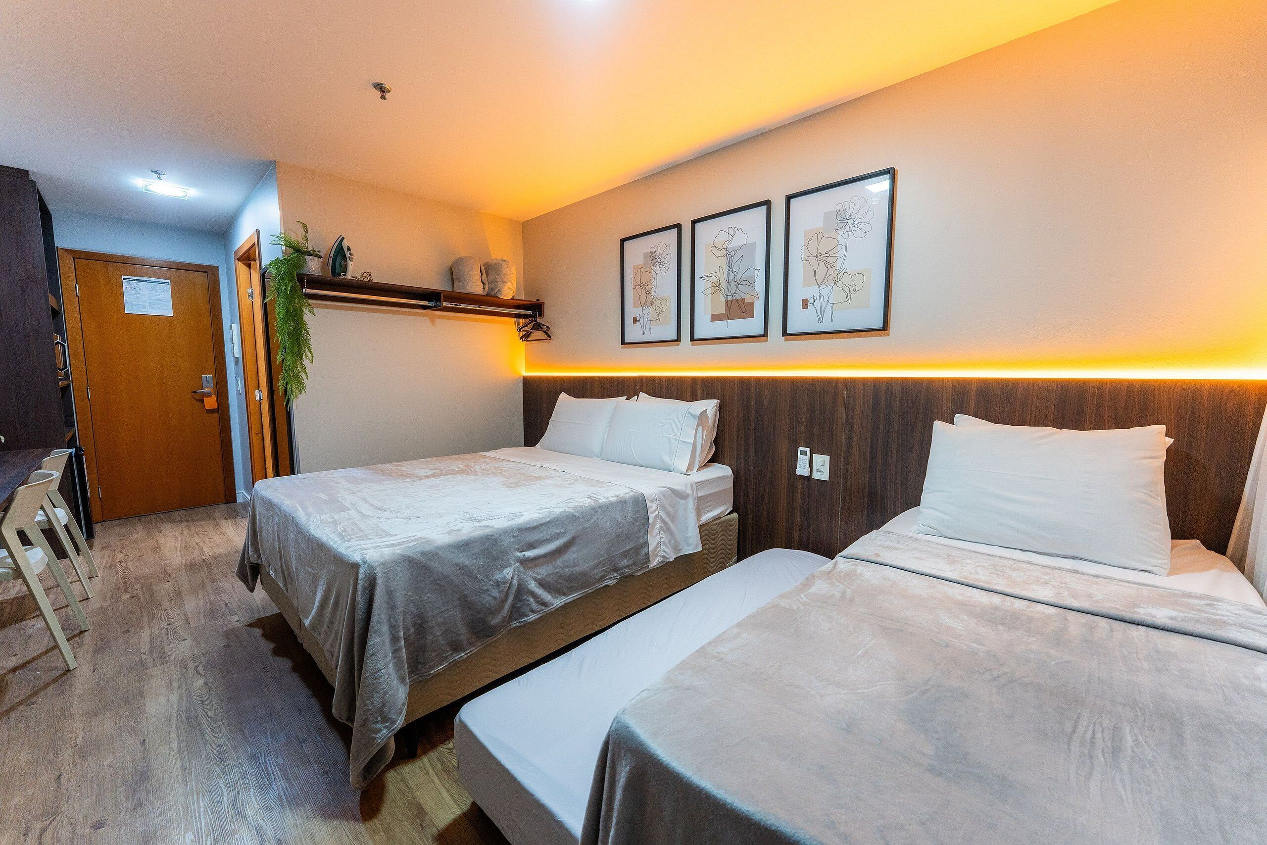 A hotel room with two beds, a desk, and a chair. There are framed pictures on the wall and a plant on the desk.