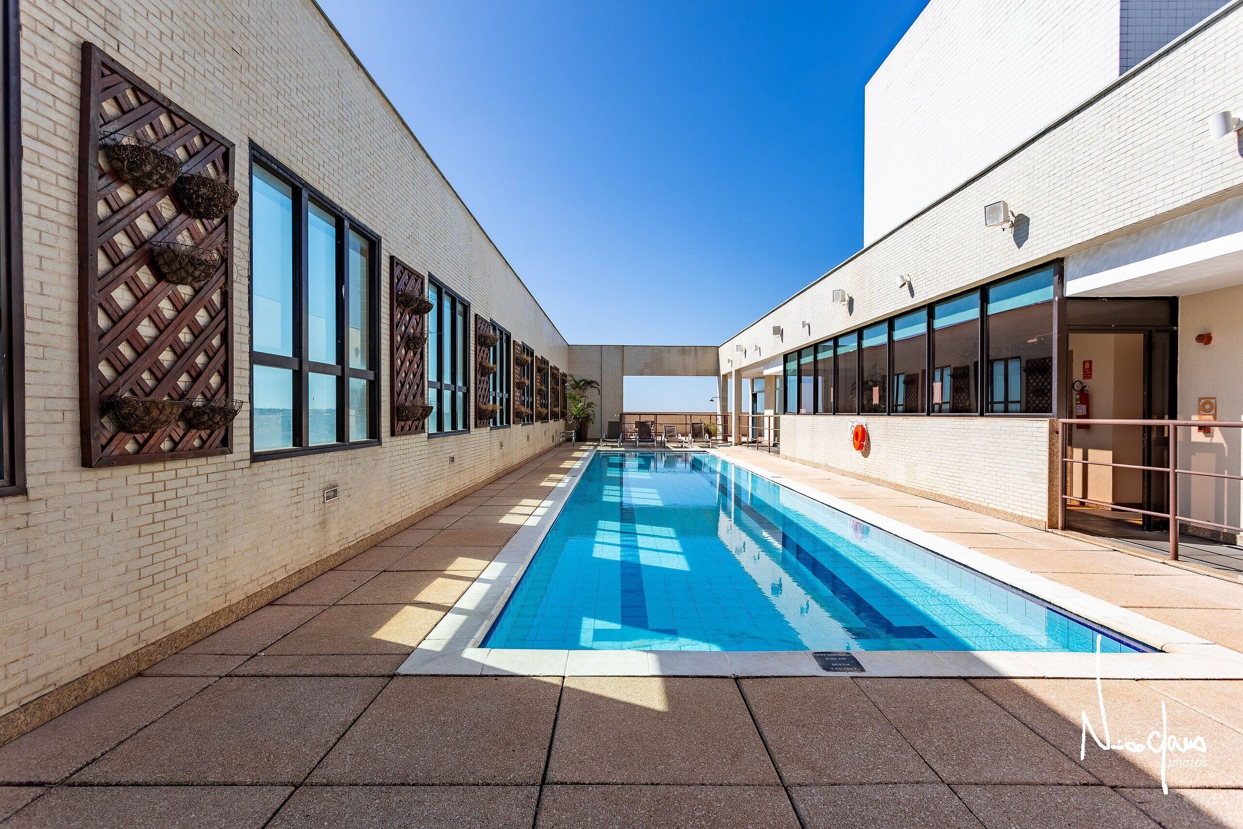 A swimming pool with a clear blue water surface, surrounded by a brick wall and large windows on one side, and a white building on the other.